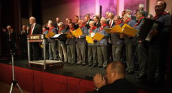 Kooperation mit Musik: Doug Armitage (vorn links), Vorsitzender des K Shoes Choir aus Kendal, präsentiert seine Mitgkieder in schwarzen Jacken und den VCR-Männerchor aus Rinteln, der in Fischerhemden Shanties sang, beim Finale im Kendaler Rathaussaal. Foto: dil